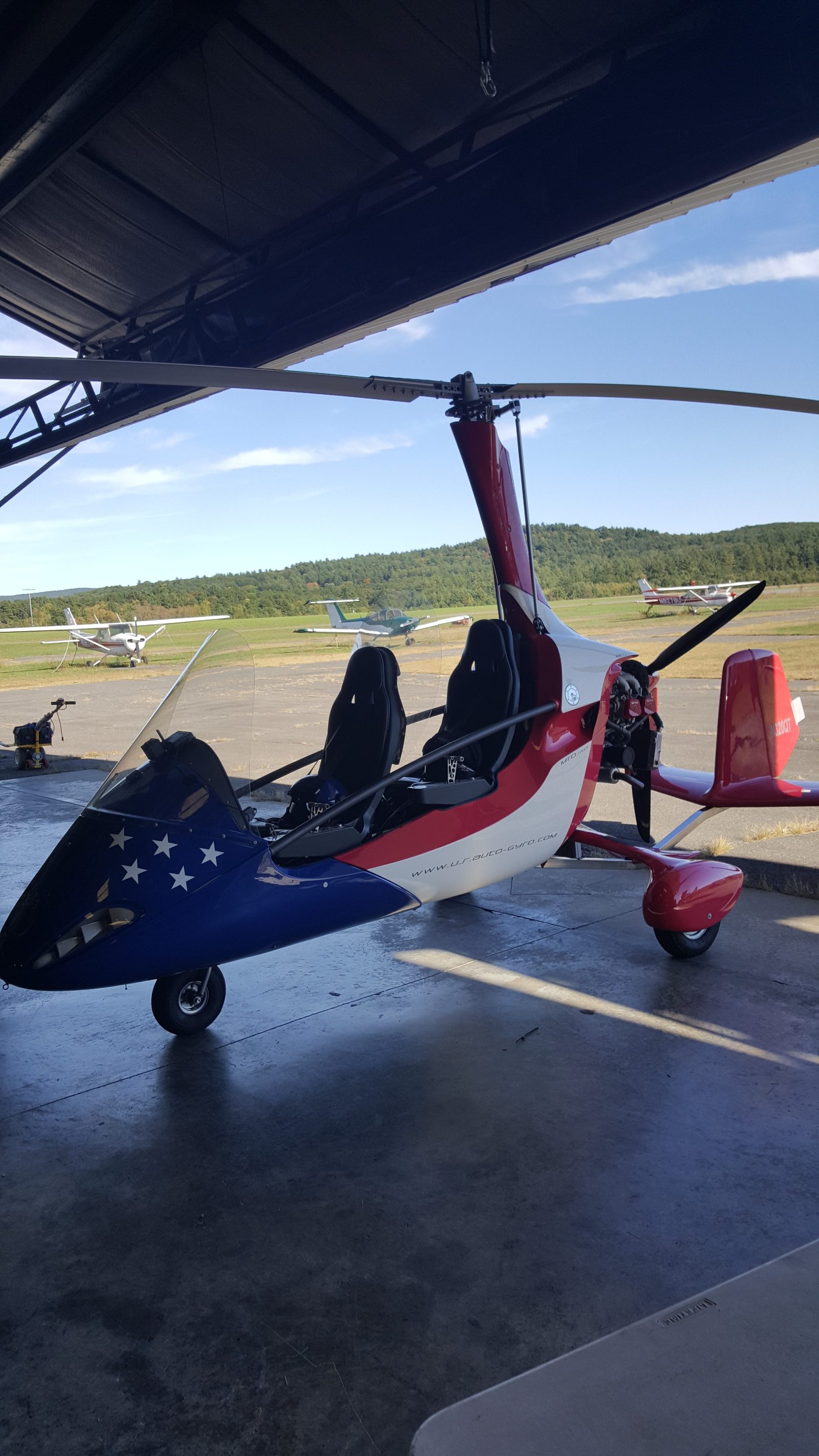Red, white and blue gyrocopter parked under a hangar canopy